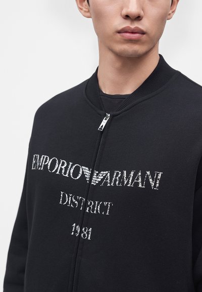 Homme portant une veste zippée noire avec un texte blanc "EMPORIO ARMANI DISTRICT 1981" sur le devant et un col côtelé.