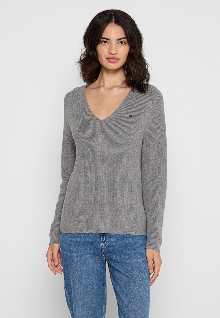 Grauer gerippter Pullover mit tiefem V-Ausschnitt und langen Ärmeln, versehen mit einem kleinen Logo auf der Brust. Kombiniert mit blauen Jeans.
