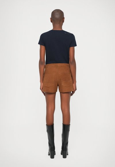 Shorts en daim marron avec deux poches arrière, associés à un t-shirt ajusté bleu marine. Le modèle porte des bottes à talons noires.