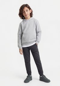 Child with shoulder-length brown hair wearing a light gray hoodie, white undershirt, dark gray pants, and dark gray sneakers standing on white background.