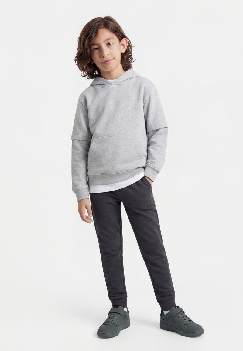 Child with shoulder-length brown hair wearing a light gray hoodie, white undershirt, dark gray pants, and dark gray sneakers standing on white background.