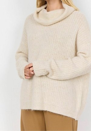 Žena v krémovém hrubém pleteném roláku s dlouhými rukávy a kamelovými kalhotami.