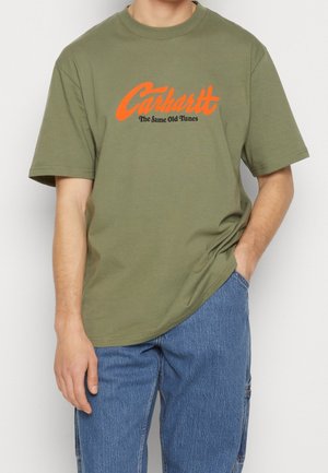 Man draagt een olijfgroen Carhartt t-shirt met oranje en zwarte tekst "The Same Old Tunes", gecombineerd met een blauwe spijkerbroek, hand in de zak.