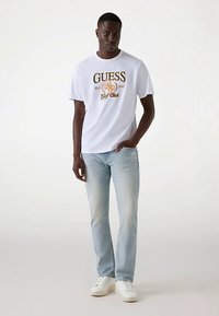 T-shirt blanc à manches courtes avec un logo "GUESS Golf Club" en or et noir, associé à un jean bleu clair et des baskets blanches.