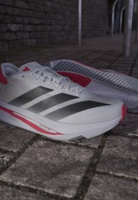 Hvide og røde Adidas løbesko med sorte striber placeret på en stenbelægning mod en sløret murstensvæg i baggrunden.