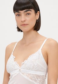 Top de lenjerie alb cu dantelă la margini, cu bretele ajustabile, având modele florale și un buton decorativ mic în mijlocul frontului.