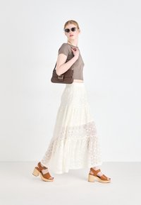 Top castanho curto, saia maxi de renda em tom marfim, sandálias de salto castanhas com saltos de madeira robustos e óculos de sol escuros, contra um fundo neutro.