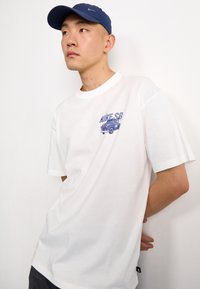 Bílá tričko s krátkým rukávem a kulatým výstřihem, s modrou grafikou vintage kamionu a textem "NIKE SB". Měkký textilní materiál.