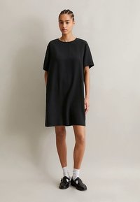 Žena stojaca v voľných čiernych šatách s krátkym rukávom, bielych ponožkách a čiernych topánkach na pozadí jednofarbného svetlého pozadia.