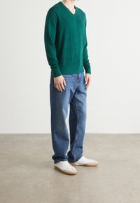 Maglione verde in lana a scollo a V, vestibilità rilassata, abbinato a jeans denim blu. Sneakers bianche con suola in gomma, in piedi su un pavimento neutro.