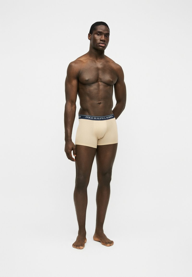 Homme musclé debout pieds nus portant un boxer beige avec une ceinture bleu marine marqué « Polo Ralph Lauren » sur un fond blanc uni.