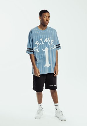 Blue striped oversized shirt with white graphic and number one, paired with black shorts featuring white logo, white sneakers, and patterned socks.