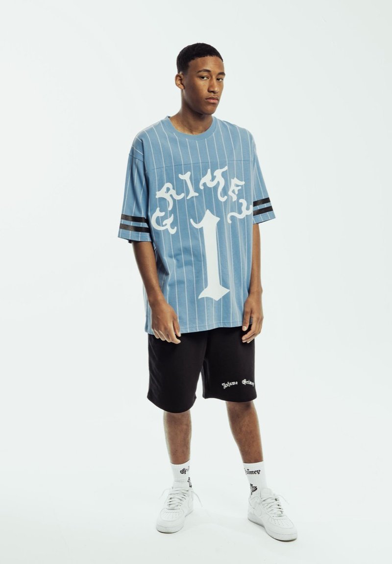 Camisa azul a rayas oversize con gráfico blanco y el número uno, combinada con pantalones cortos negros con logo blanco, zapatillas blancas y calcetines con estampado.