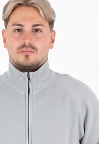 Giacca zip-up grigia chiara con colletto alto, texture a costine e dettagli in zip nera, caratterizzata da un design aderente e senza motivi visibili.