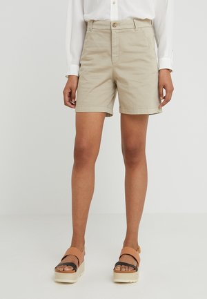 Femme portant un short beige longueur genou, une chemise blanche à manches longues et des sandales compensées couleur beige avec des brides noires aux orteils.