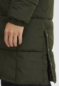 Oliven gr�øn polstret jakke med en tekstureret overflade, elastiske manchetter og trykknapper på siden, med et nærbillede af en hånd på stoffet.