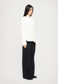 Cămașă albă cu mânecă lungă și bătaie la gât, purtată cu pantaloni largi, de culoare bleumarin închis și pantofi negri, expusă pe un fundal simplu.