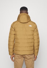 Brun puffjacka med huva, med horisontella quiltade sektioner och en liten vit logotyp på övre ryggen.