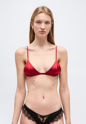 Młoda kobieta o długich blond włosach, w czerwonym satynowym biustonoszu i czarnych majtkach z koronkowymi wykończeniami, stojąca na tle jednolitego, jasnego tła.