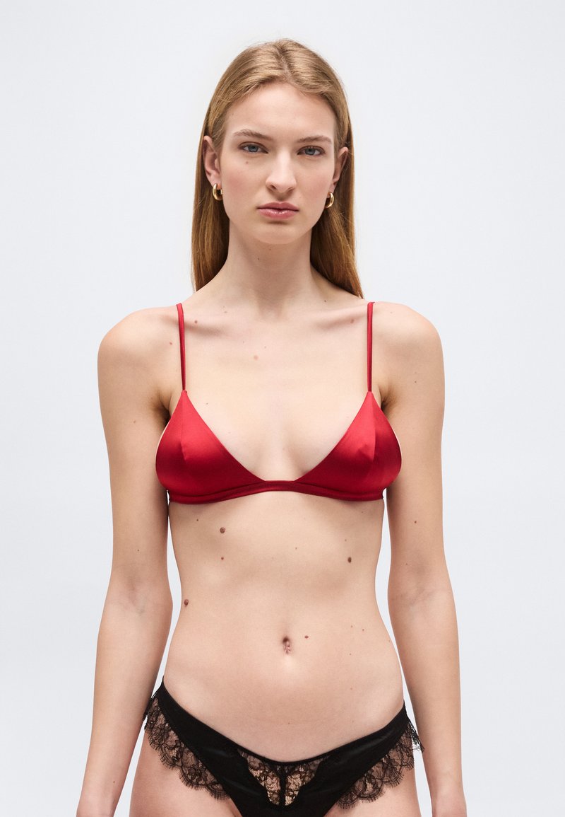 Νεαρή γυναίκα με μακριά ξανθά μαλλιά, φοράει κόκκινο σατέν σουτιέν και μαύρα εσώρουχα με δαντέλα, στέκεται μπροστά σε απλό φωτεινό φόντο.