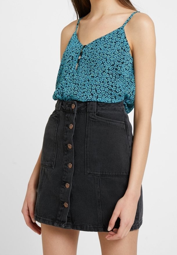 Femme portant un haut sans manches bleu à motifs floraux, rentré dans une jupe en jean noire taille haute avec des boutons sur le devant et des poches à l'avant.