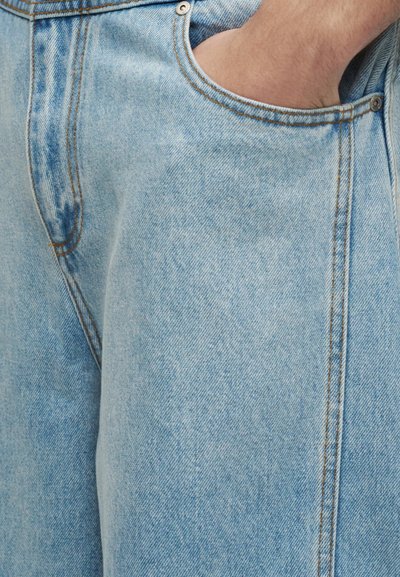 Κοντινή λήψη ενός χεριού που έχει μπει στην εμπρόσθια τσέπη ενός ανοιχτόχρωμου τζιν με εμφανή ραφές και μεταλλικό καινό.