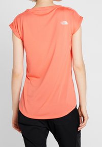 Dámské tričko s krátkým rukávem v korálové barvě, vyrobené z hladké tkaniny. Má kulatý výstřih a malé bílé logo na horní části zad.