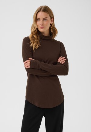 Femme aux longs cheveux blonds porte un col roulé marron et un pantalon noir, debout, les bras croisés, regardant légèrement sur le côté, sur un fond uni.