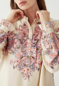 Cremefarvet bluse med lange ærmer, der har et blomstret paisley-mønster i pink, blå og brun. Rynket front og knapdetaljer ved ærmekanterne.