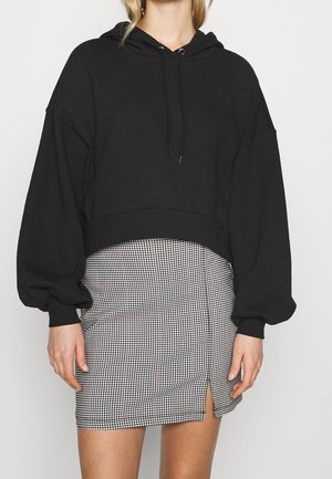Kobieta w czarnej, krótkiej bluzie z kapturem i sznureczkami oraz czarno-białej mini spódnicy w kratę z małym rozcięciem z przodu.