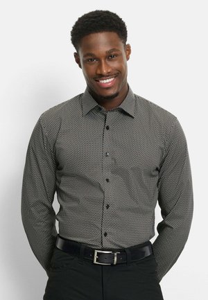 Lächelnder Mann mit kurzen lockigen Haaren, der ein schwarz-weiß gemustertes Langarmshirt und schwarze Hose mit Gürtel trägt, vor weißem Hintergrund stehend.