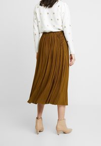 Falda plisada de longitud media en tejido marrón intenso, con cintura elástica. Combinada con un suéter blanco con puntos dorados y botas de tobillo beige.