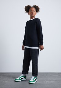 Svart langermet genser med hvit krage, kombinert med oppbrettede mørke jeans og grønne, svarte og hvite sneakers. Betongbakgrunn.