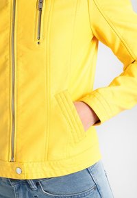 Giacca di pelle gialla con zip, tasche laterali e dettagli di cucitura visibili. Il design è aderente con una lunghezza cropped.