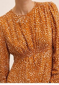 Femme portant une robe orange rouille avec des taches blanches abstraites, taille élastique froncée, manches longues et encolure ronde.