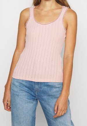 Kobieta w jasno różowym bezrękawnym dzianinowym topie z falbanką i jasnoniebieskich dżinsach, stojąca na jednolitym tle.