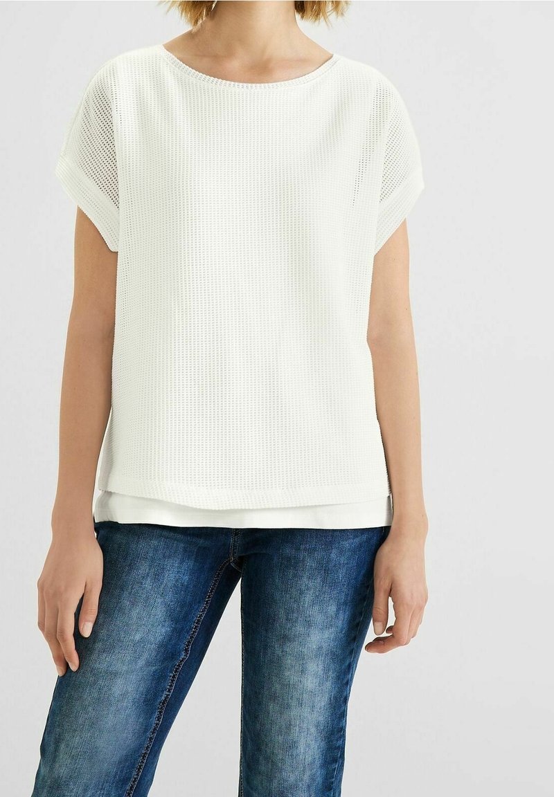 Vit kortärmad topp med ett texturerat mesh-mönster och en vid halsringning, lager på lager över ett slätt, vitt innerskikt. Tillsammans med blå jeans.