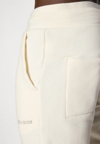 Beige joggers i bomuldsblanding med sidelommer, udstyret med en syet søm og diskret branding på venstre ben. Blød tekstur, afslappet pasform.