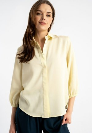 Kobieta o długich brązowych włosach, ubrana w luźną, bladożółtą bluzkę zapinaną na guziki i ciemne spodnie na jednolitym białym tle.