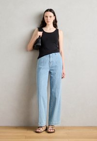 Black sleeveless top, light blue high-waisted wide-leg jeans, and brown sandals. Model holds a small black bag; background is neutral.