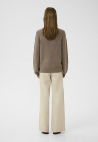 Brauner Wollpullover mit lockerer Passform, überschnittenen Schultern und geripptem Saum, kombiniert mit beigen, weiten Hosen und schwarzem Schuhwerk.