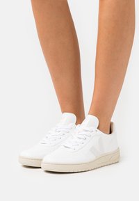 Hvide lædersneakers med en struktureret overflade, der har flade hvide snørebånd, en kontrastfarvet beige sål og et logo på hælen.