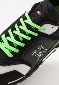 Černá sportovní bota se zelenými tkaničkami, texturovaný černobílý materiál, detail loga a gumová podešev s náznakem neonově zelených akcentů.