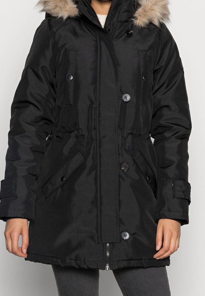 Parka noire avec une capuche en fausse fourrure, dotée d'une fermeture éclair à l'avant, de boutons-pression et de deux poches latérales. Le tissu a une finition lisse et brillante.