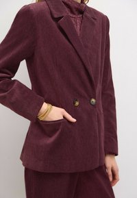 Burgundy corduroy kaksirivinen bleiseri, jossa on herringbone-kudos, kaksi etunappia ja sivutaskuyksityiskohta. Pitkät hihat.