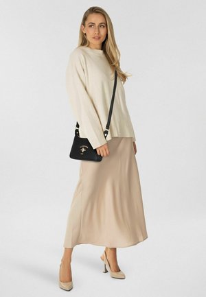 Donna che indossa un maglione beige e una gonna, tacchi beige, tiene una borsa a tracolla nera con logo dorato, in piedi davanti a uno sfondo neutro.