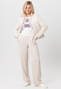 Lichtgewicht, gewatteerde crèmekleurige jas met roze afwerking, gedragen over een witte graphic t-shirt, gecombineerd met loszittende beige broek en witte sneakers.