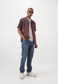 Chemise à manches courtes, bordeaux, à boutons, tee-shirt beige clair, jeans bleus, baskets blanches avec une semelle texturée, et lunettes de soleil noires.