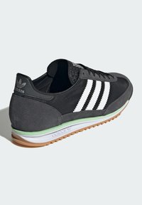 Zapatillas negras con parte superior de ante y textil, tres franjas blancas, acento verde en la entresuela y suela de goma.
