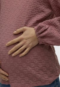 Ružičasta bluzica sa teksturom i cvjetnim uzorcima; elastični manžeti, opušteni kroj. Ruka odmara na zaobljenom trbuhu. Jednostavan dizajn s mekanim materijalom.